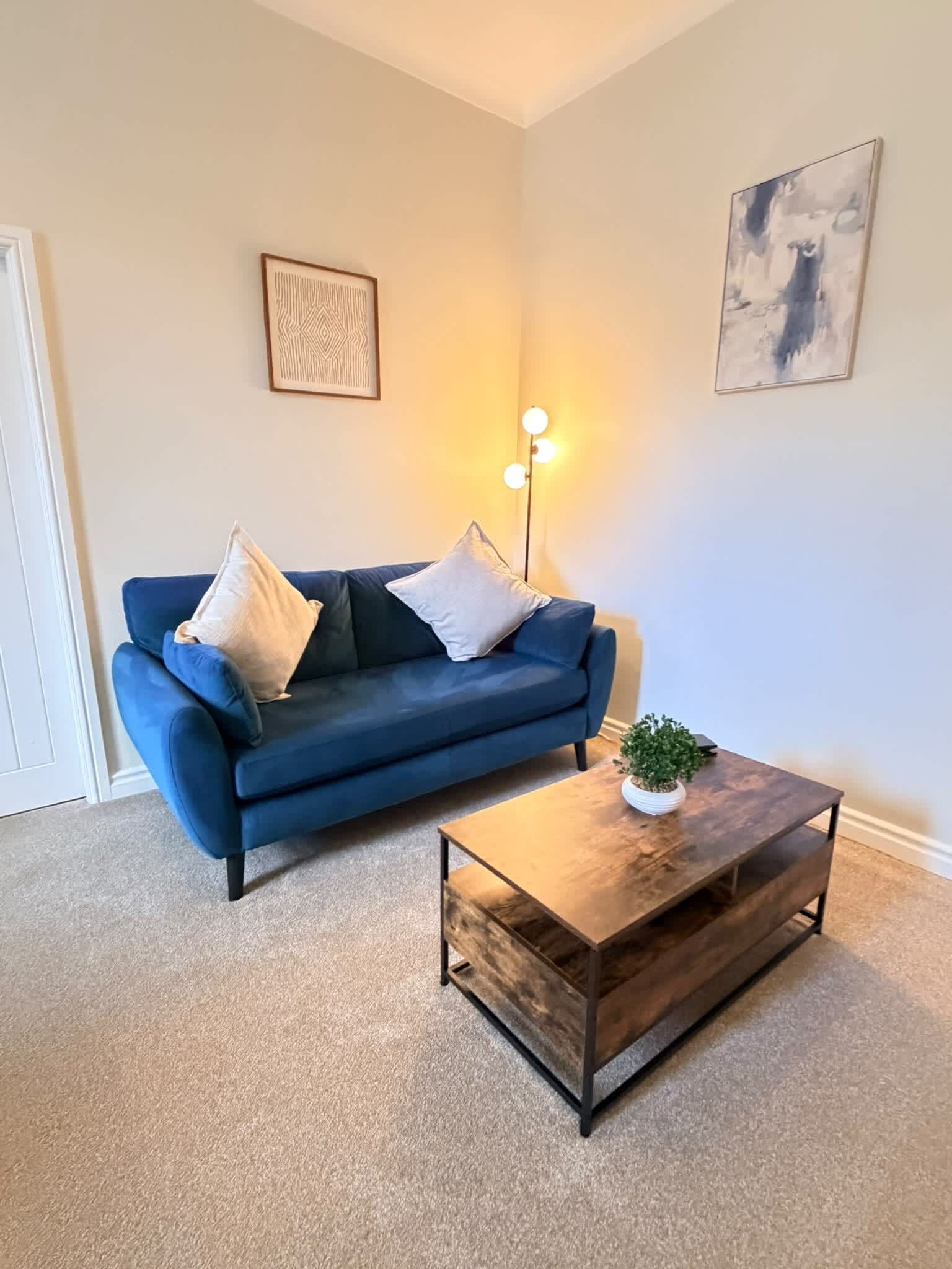 Una sala de estar con un sofá azul, dos almohadas, una mesa de centro con una planta y dos lámparas fijadas a la pared.