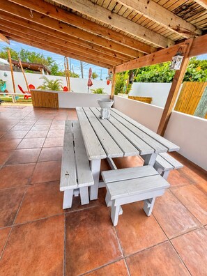 Een terras buiten met houten picknicktafels en bankjes, omgeven door een witte muur en beplanting.