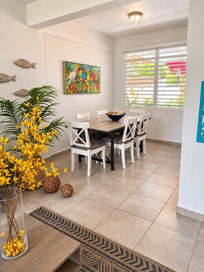 A dining area with a table and chairs, a painting on the wall, and a window with blinds. - Ramey Base 3BR/2BA / Surf + Solar + Starlink + AC (Aguadilla)
