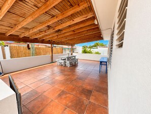 A covered patio with a tiled floor, wooden ceiling beams, and a white wall with a window. - Ramey Base 3BR/2BA / Surf + Solar + Starlink + AC (Aguadilla)