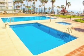 A rectangular swimming pool with a tiled deck, surrounded by palm trees and a building in the background. - Aloha Playa Apartment with a Pool and Ocean View (Benalmádena Costa)
