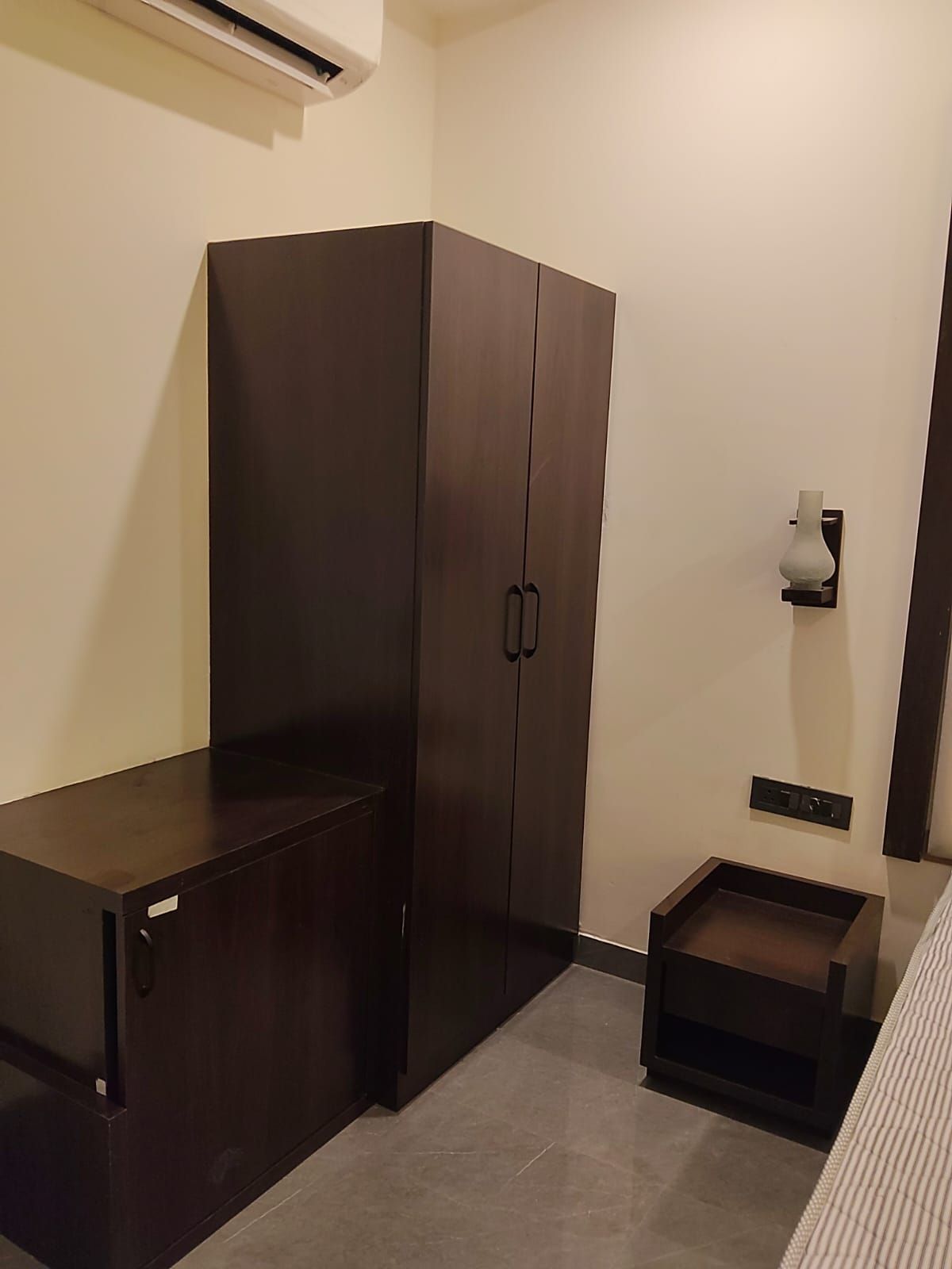 Habitació amb un armari de fusta fosca, una petita taula de nit i una prestatgeria penjada a la paret.