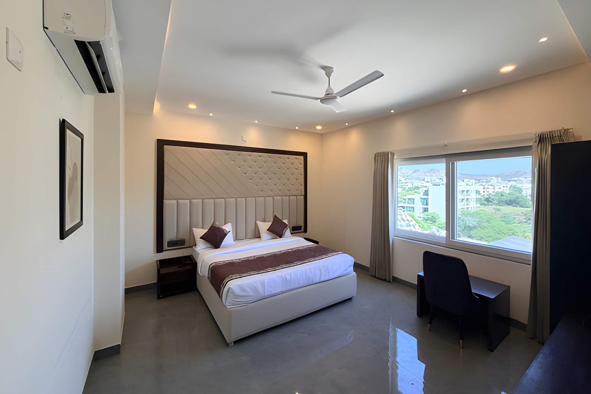 Una habitación de hotel moderna con una cama grande, un escritorio, una silla, un ventilador de techo y una ventana con vista a la ciudad.