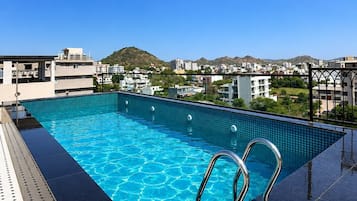 A rooftop swimming pool with clear blue water, surrounded by a black tiled deck and metal pool ladders.