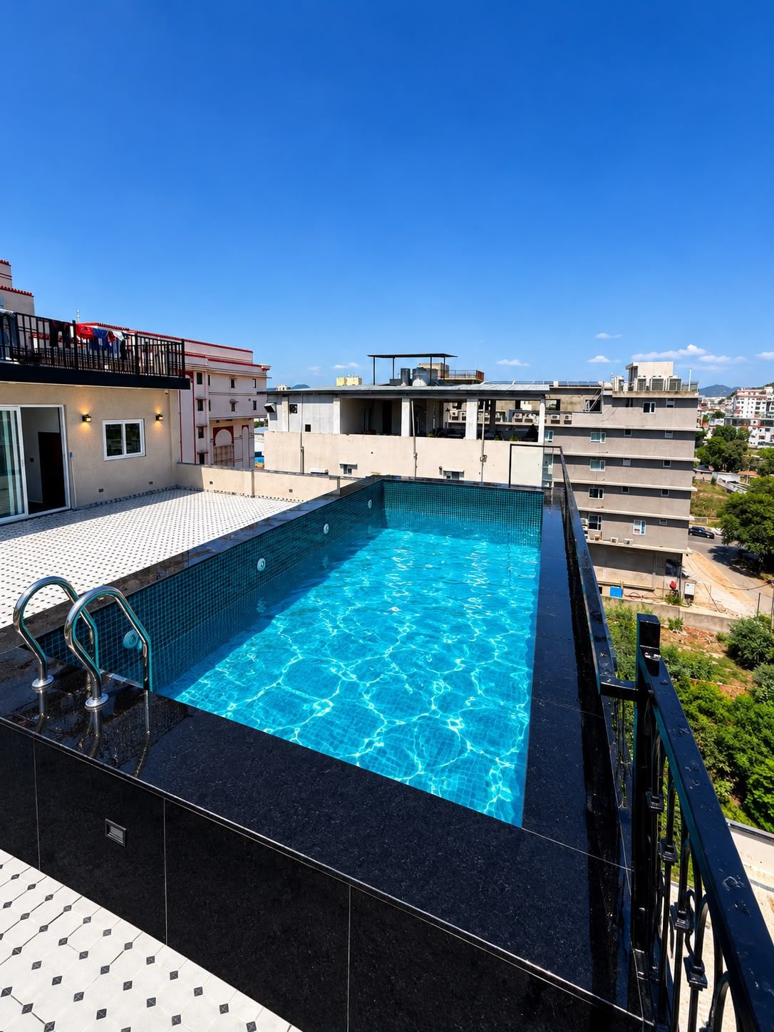 A rooftop swimming pool with clear blue water, surrounded by a black pool deck and a metal railing.