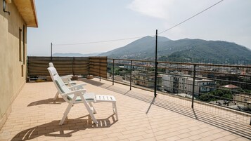 A balcony with two white lounge chairs, a small table, and a view of a cityscape and mountains in the background.