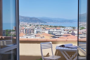 Uma varanda com vista para uma cidade costeira, uma cadeira e uma mesa com um livro.