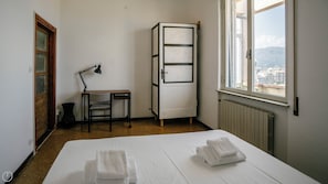 Quarto de hotel com uma cama, uma escrivaninha com cadeira, um radiador e uma janela com vista para edifícios e montanhas.