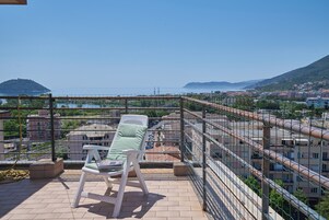 A balcony with a white chair, a view of the sea, and buildings in the background.