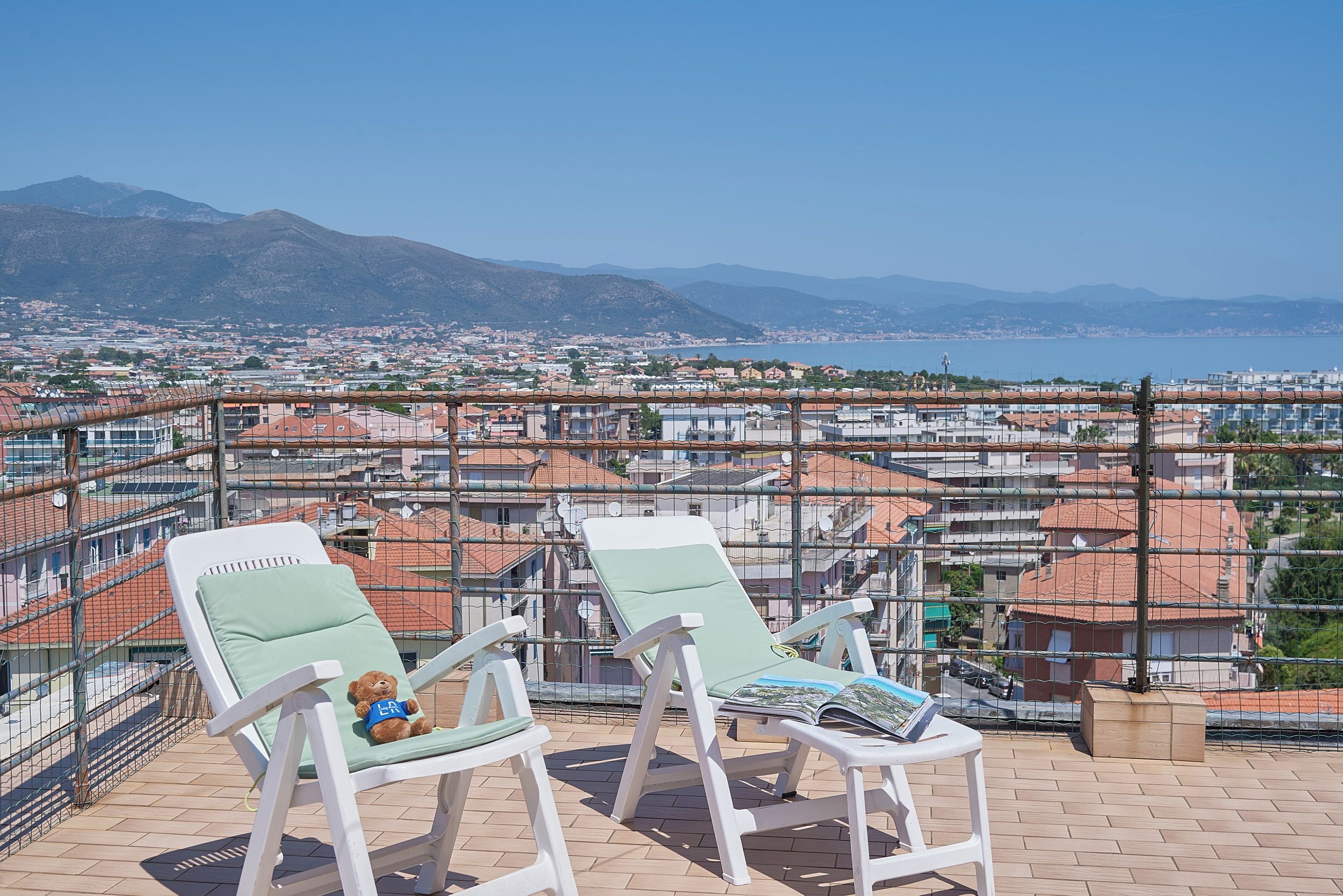 A balcony with two lounge chairs, one with a teddy bear and a magazine, overlooking a coastal cityscape with mountains in the distance.