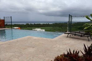 Piscina com vista para o mar, área cercada com espreguiçadeiras e paisagem verde ao fundo.