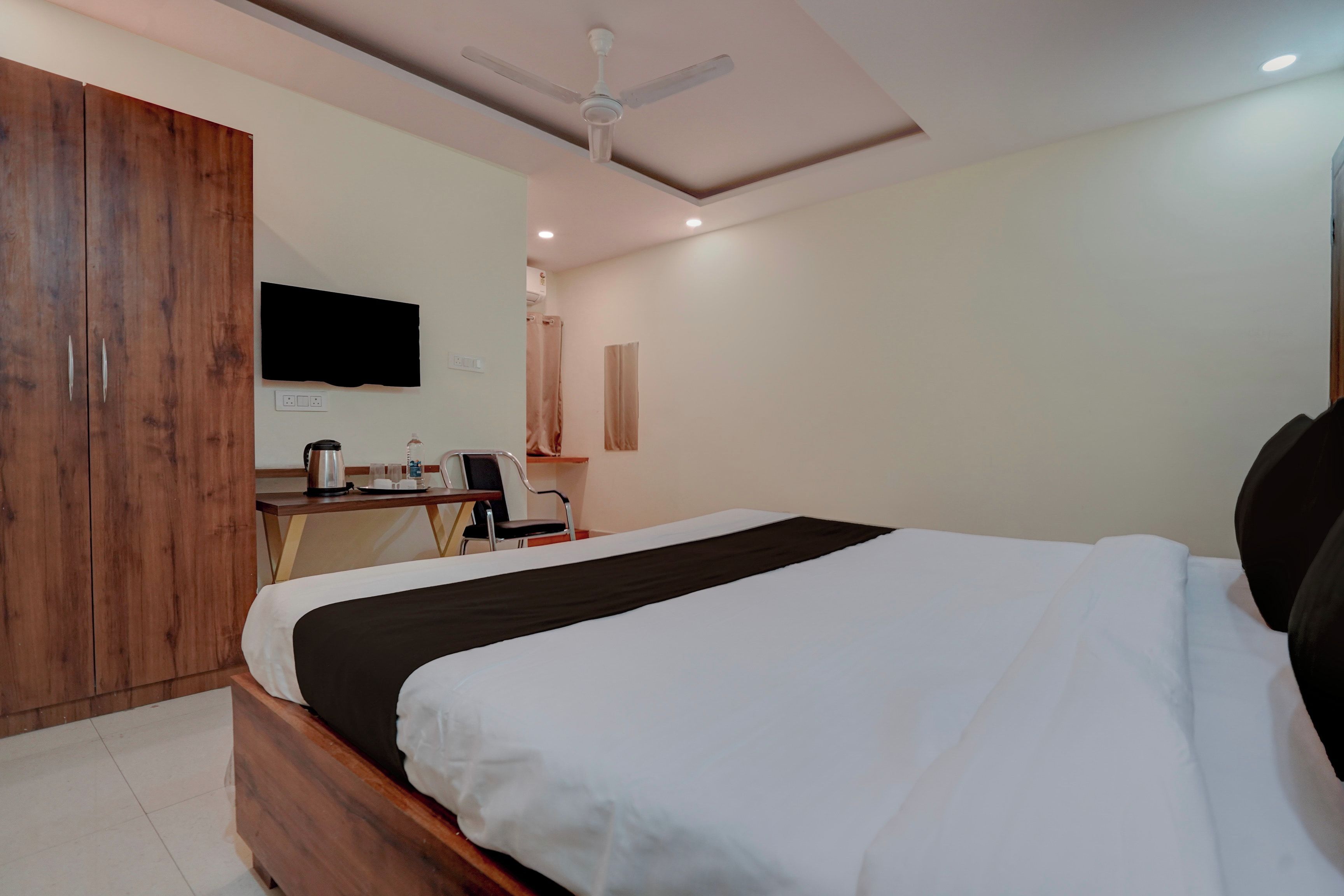 Habitación de hotel con cama, escritorio con silla, armario, ventilador de techo y televisor de pantalla plana montado en la pared.