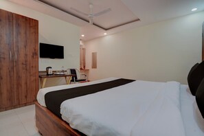 Habitación de hotel con cama, escritorio con silla, armario, ventilador de techo y televisor de pantalla plana montado en la pared.