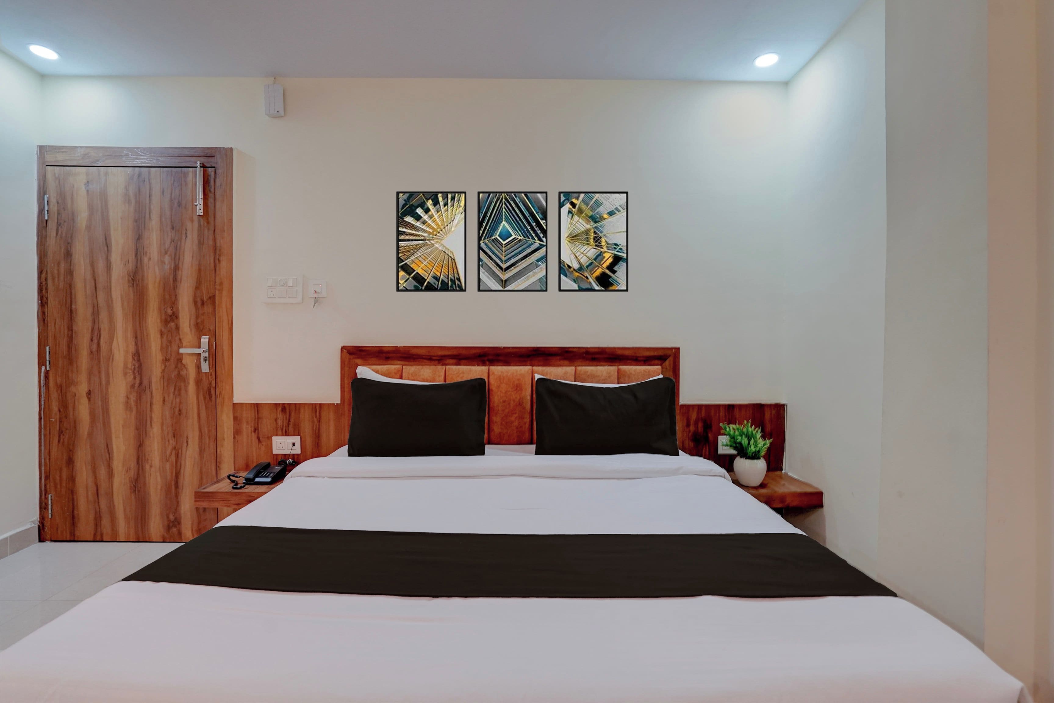 Una habitación de hotel con cabecera de madera, cama con ropa de cama blanca y negra, mesita de noche con teléfono y planta, y tres piezas de arte abstracto en la pared.