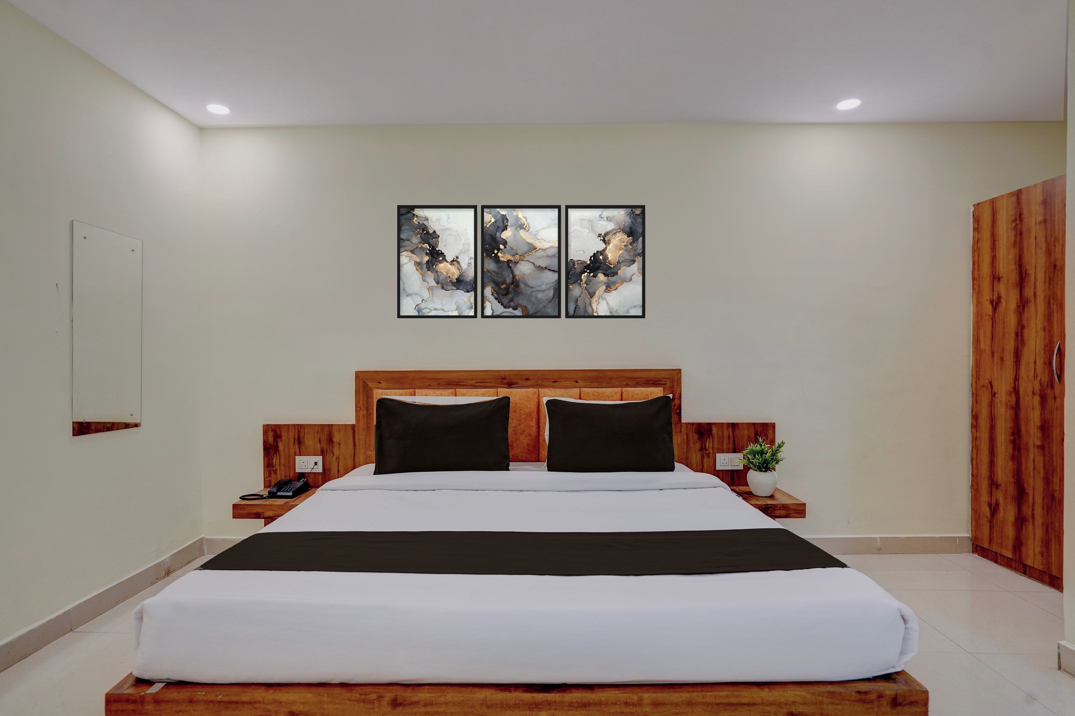 A hotel room with a large bed, wooden headboard, bedside tables, a nightstand, a telephone, a plant, and artwork on the wall.
