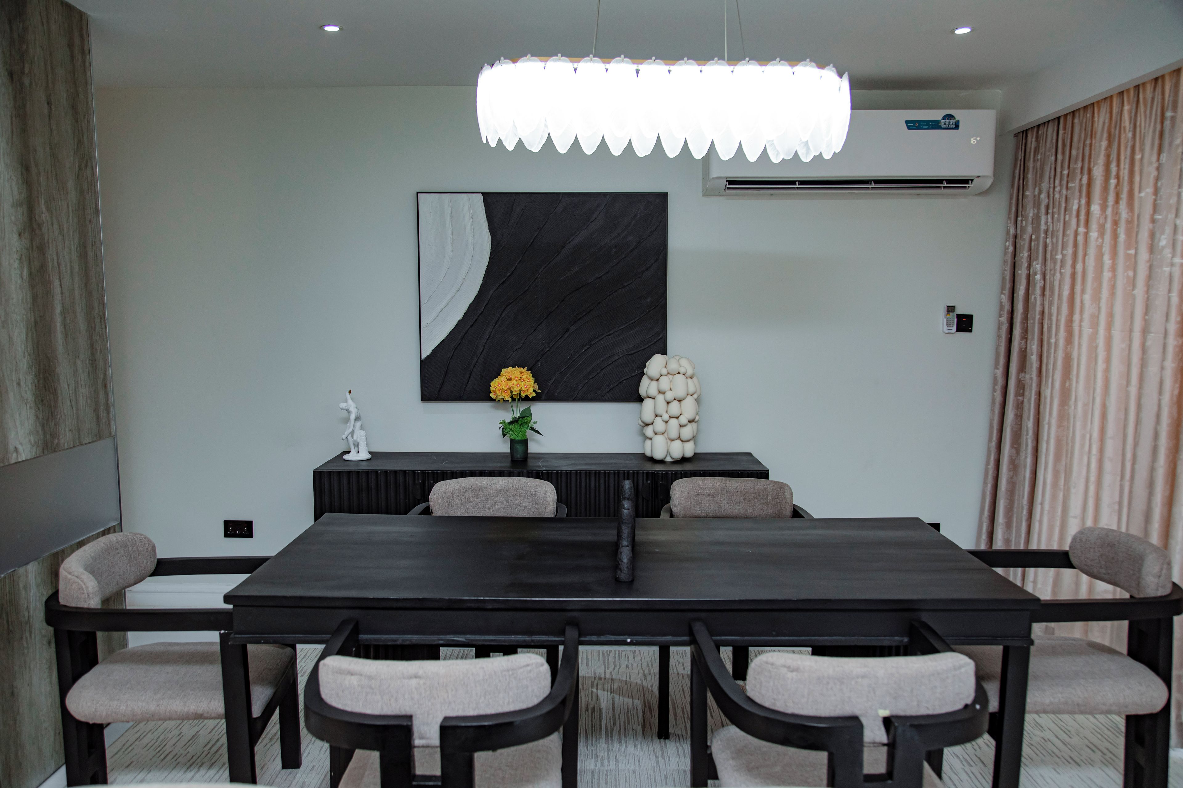 A modern conference room with a dark wooden table, six chairs, a large abstract painting, a potted plant, and a decorative white sculpture.