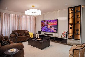 A modern living room with a large flat-screen TV, a brown sofa, a coffee table, and a decorative shelf with plants. - Villa De Fredwaa Unit 2 (Dar es Salaam)