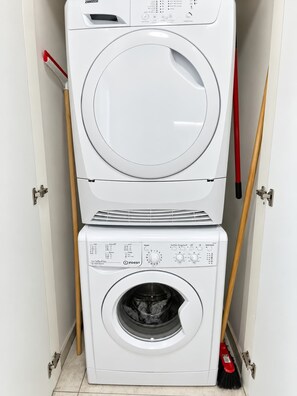 A stacked washer and dryer unit with a control panel, located in a narrow space with a door and a broom leaning against the wall. - Family Friendly Downtown Apartment with Free Parking By Isrentals (Jerusalem)