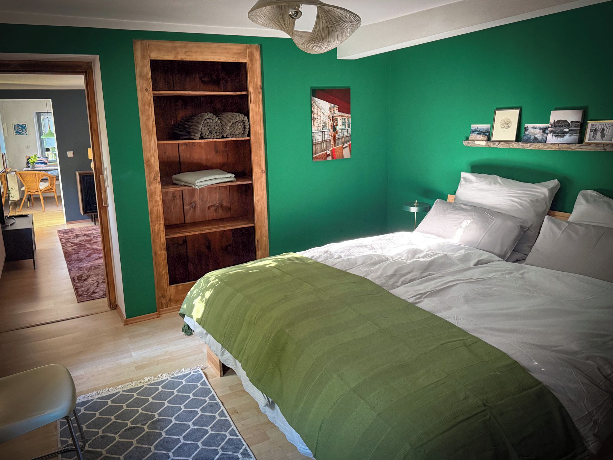 A bedroom with a green wall, a wooden wardrobe, a bed with white bedding, and a view of a cityscape through a window.