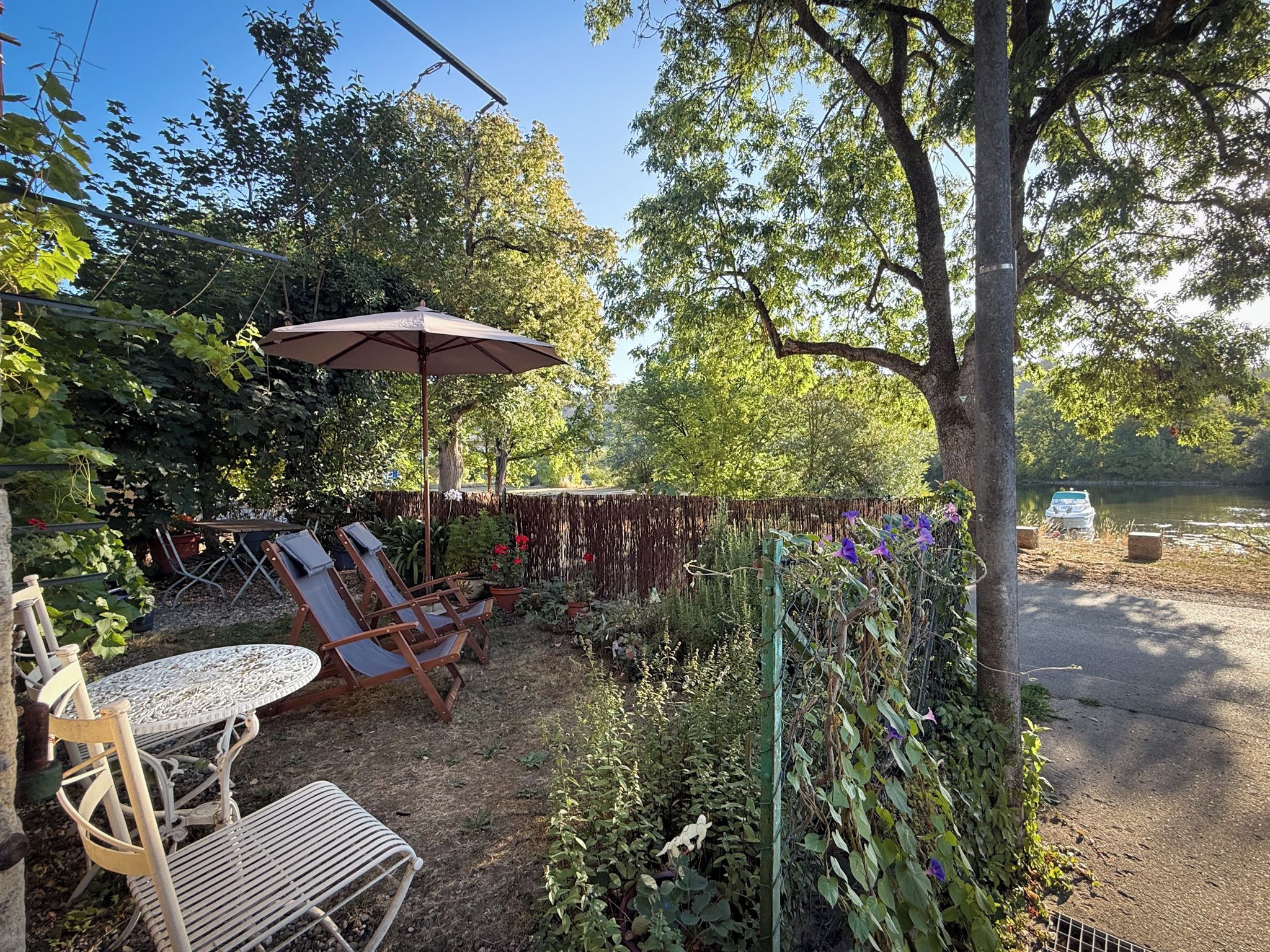 An outdoor seating area with a table and chairs, surrounded by lush greenery and a pergola with an umbrella.