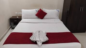 A neatly made bed with white bedding and a red accent pillow, a folded towel on the bed, a small bedside table with a remote control, and a dark wooden wardrobe.