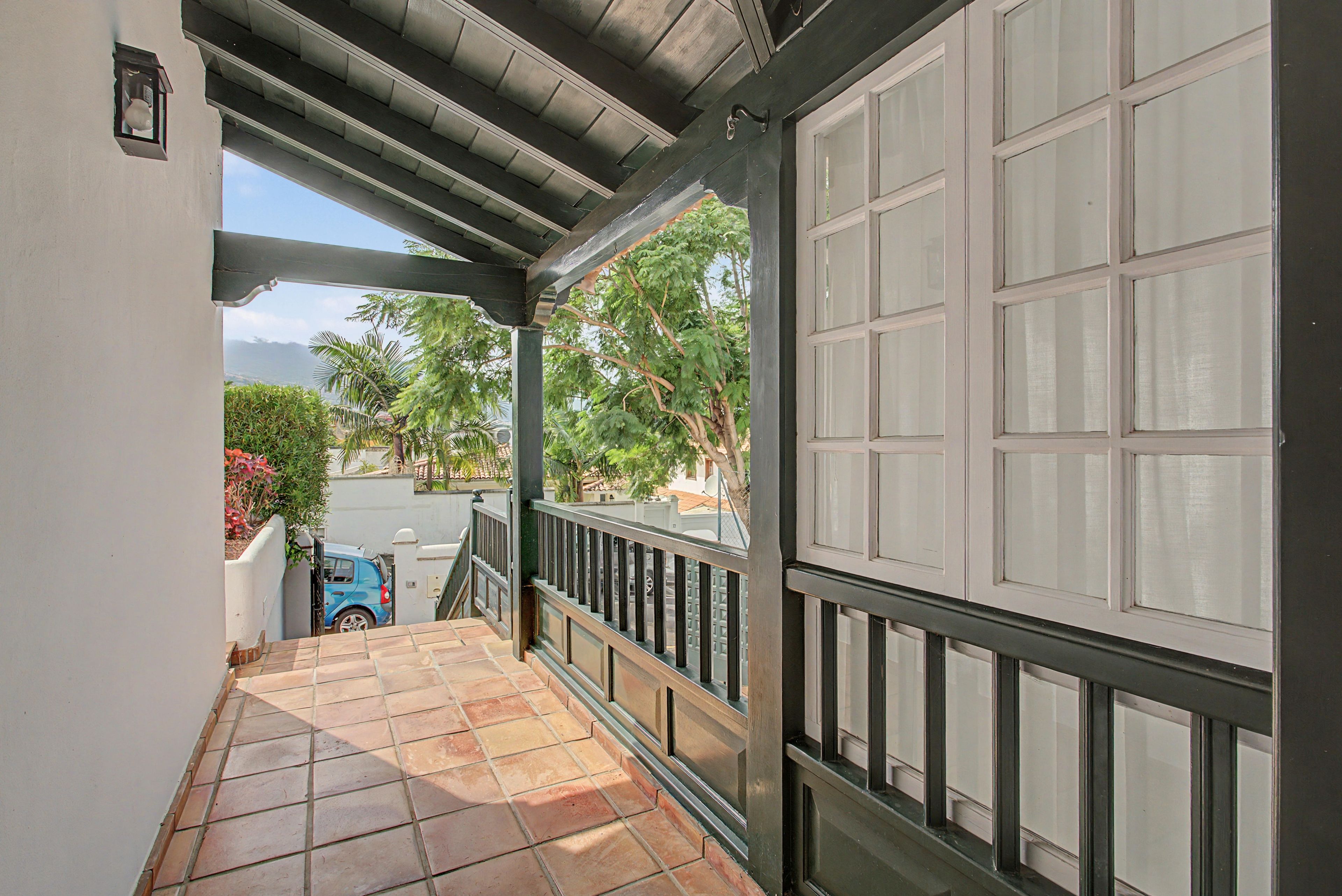 A covered balcony with a tiled floor, a metal railing, and a view of a parked car and greenery.