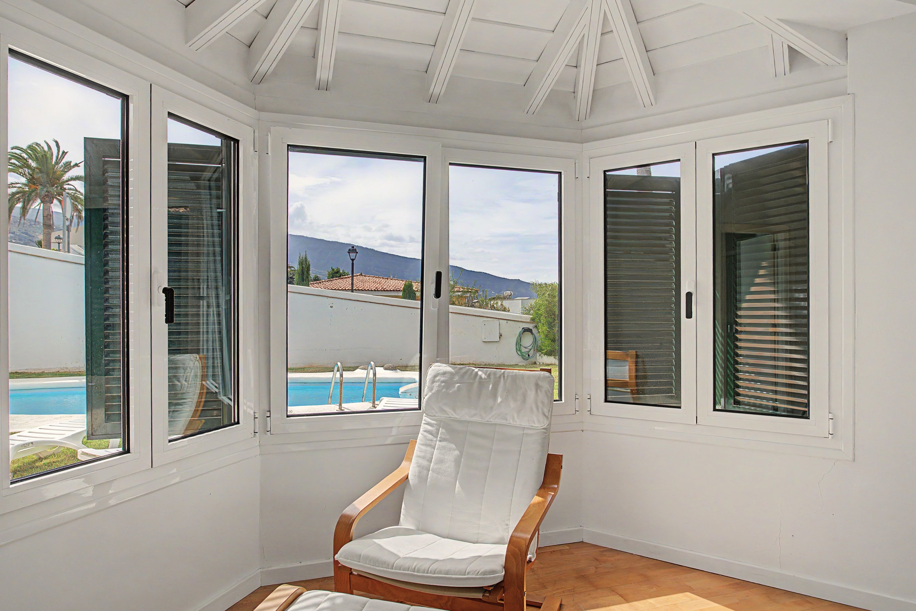 A room with a white chair, large windows, and a view of a pool and mountains.