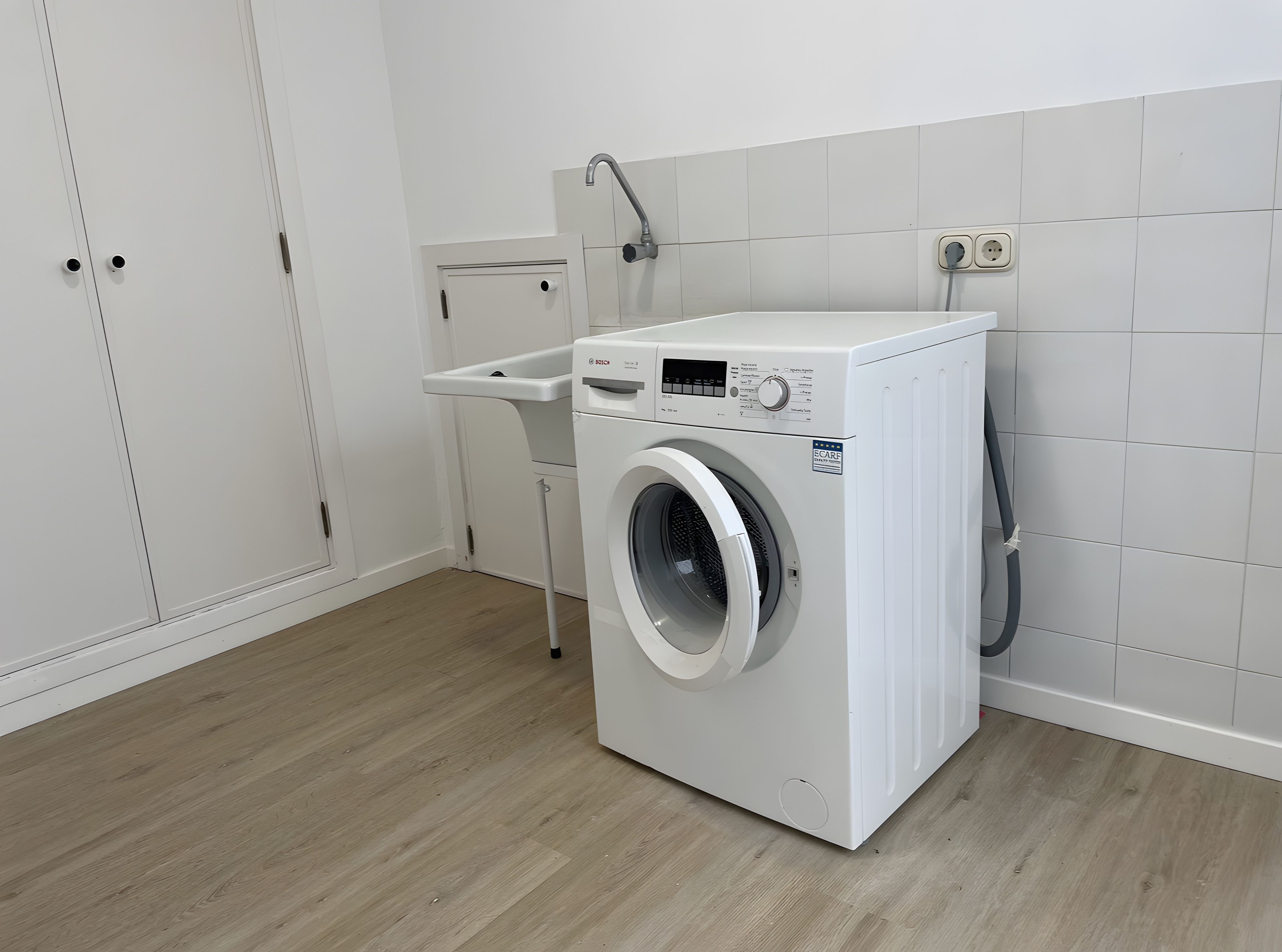 Een compacte, witte wasmachine met een frontlader in een nette, moderne wasruimte met houten vloeren en witte betegelde wanden.
