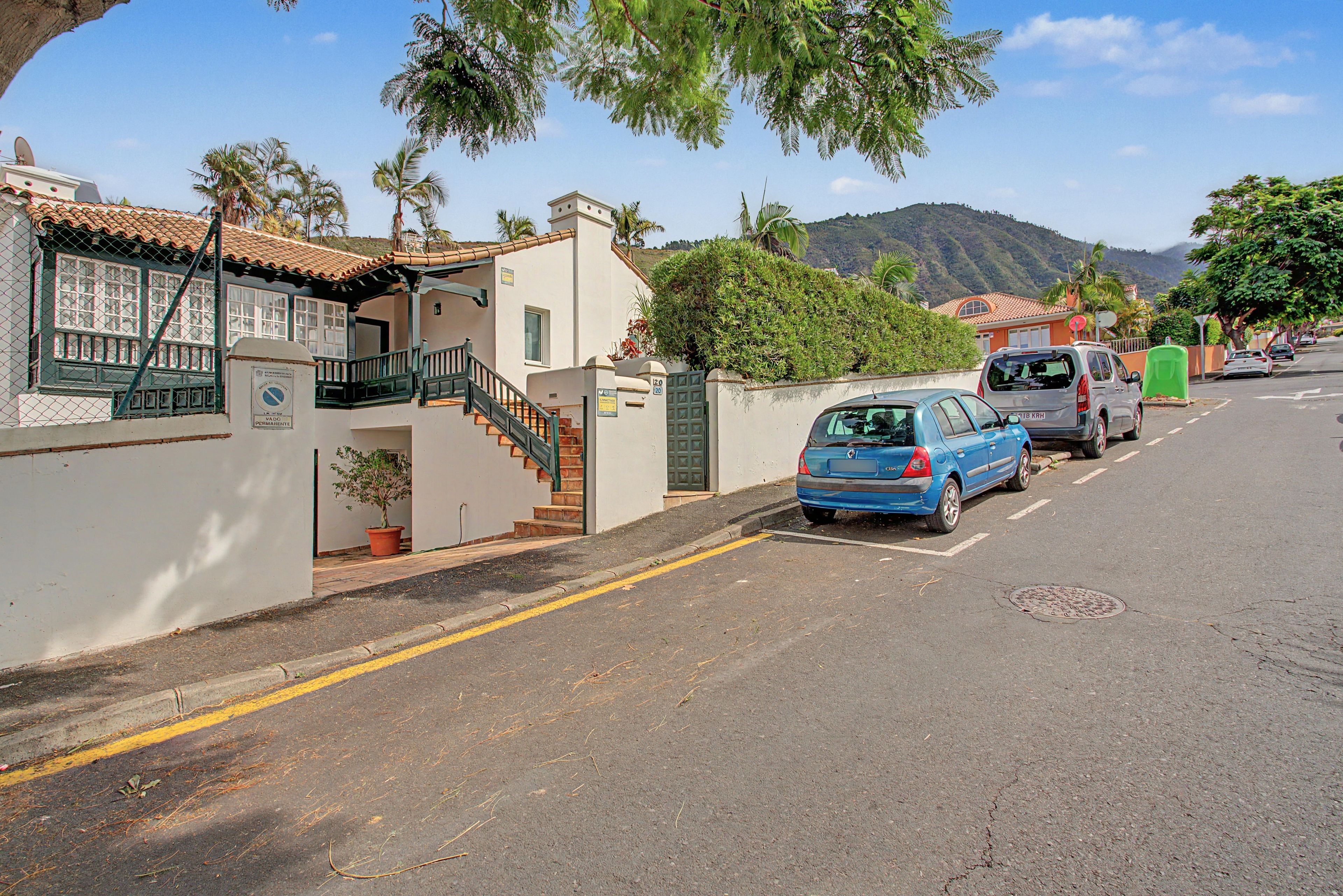 A residential street with houses, a blue car parked on the side, and a hilly landscape in the background.