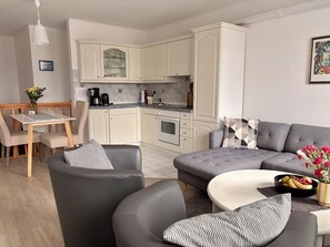 A modern kitchen with white cabinets, a dining area with a table and chairs, and a grey sofa with patterned cushions. - Fischerstuuv - 50-square-meter apartment for 2 in downtown Husum (Husum)