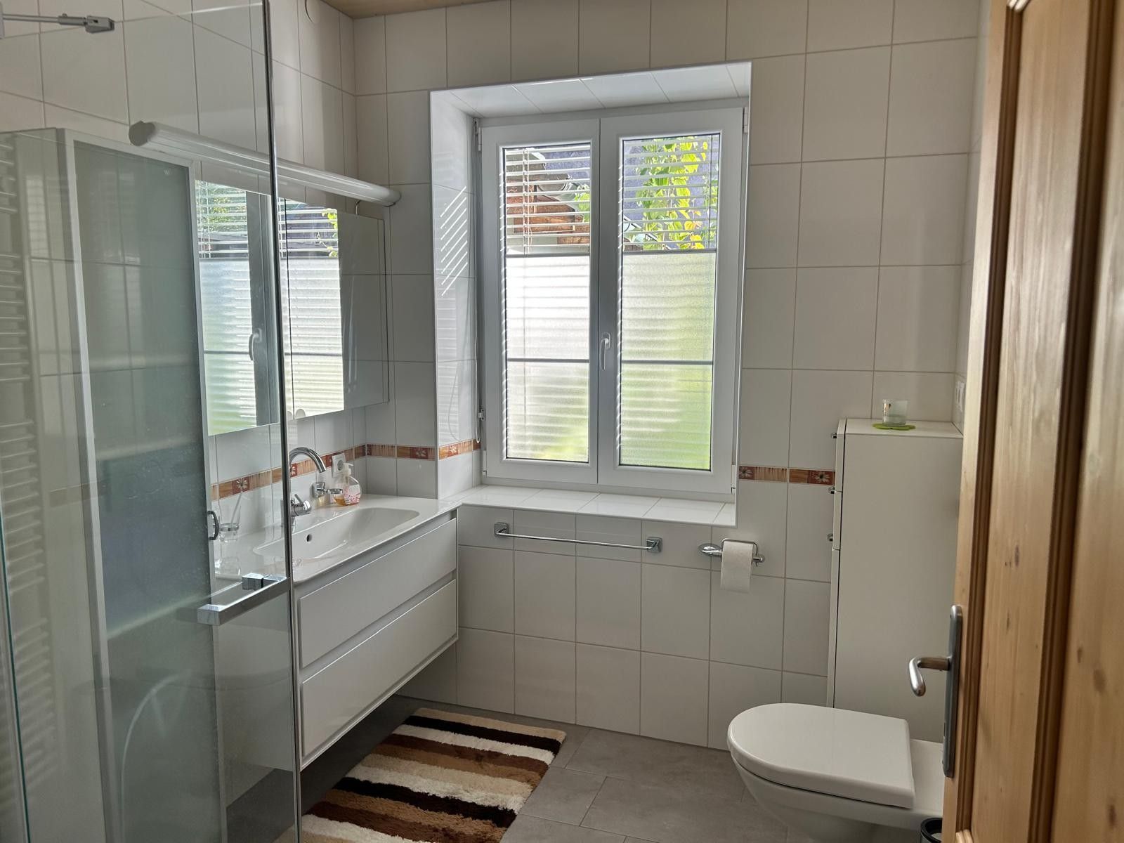 Ein Badezimmer mit Duschkabine aus Glas, einem weißen Waschbecken, einer Toilette und einem Fenster mit Jalousien.