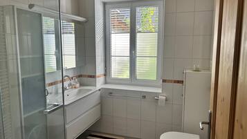 Ein Badezimmer mit Duschkabine aus Glas, einem weißen Waschbecken, einer Toilette und einem Fenster mit Jalousien.