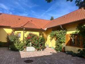 Ein gelbes Gebäude mit einem roten Ziegeldach, einem gepflasterten Innenhof und einem Blumenbeet mit rosa Blüten.