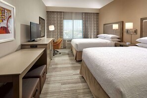 Ein Hotelzimmer mit Schreibtisch, zwei Betten, einem Sessel und einem großen Fenster mit Vorhängen.