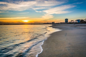 Ein Strand mit Sonnenuntergang, ruhigem Wasser und einer Stadtansicht im Hintergrund.