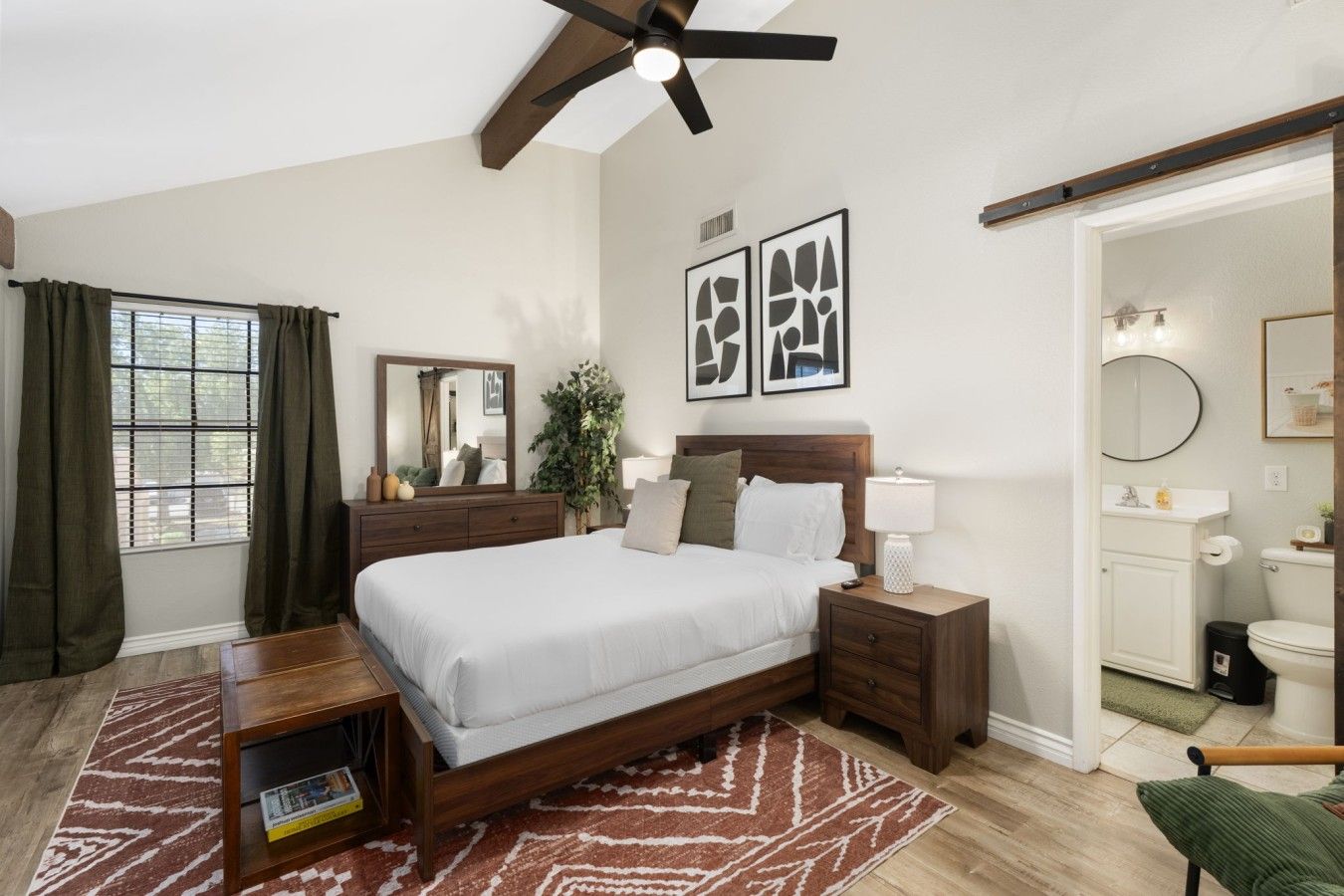 Un dormitorio con cama, mesitas de noche, espejo, ventana con cortinas, ventilador de techo y obras de arte en la pared.