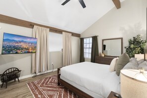 Un dormitorio con una cama grande, un televisor montado en la pared, una ventana con cortinas, una silla de madera y un espejo.