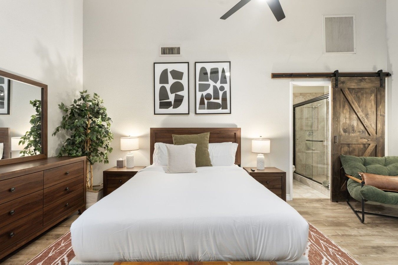 Un dormitorio con una cama grande, dos mesitas de noche con lámparas, un aparador, un espejo, una planta verde y una puerta corrediza de estilo granero.