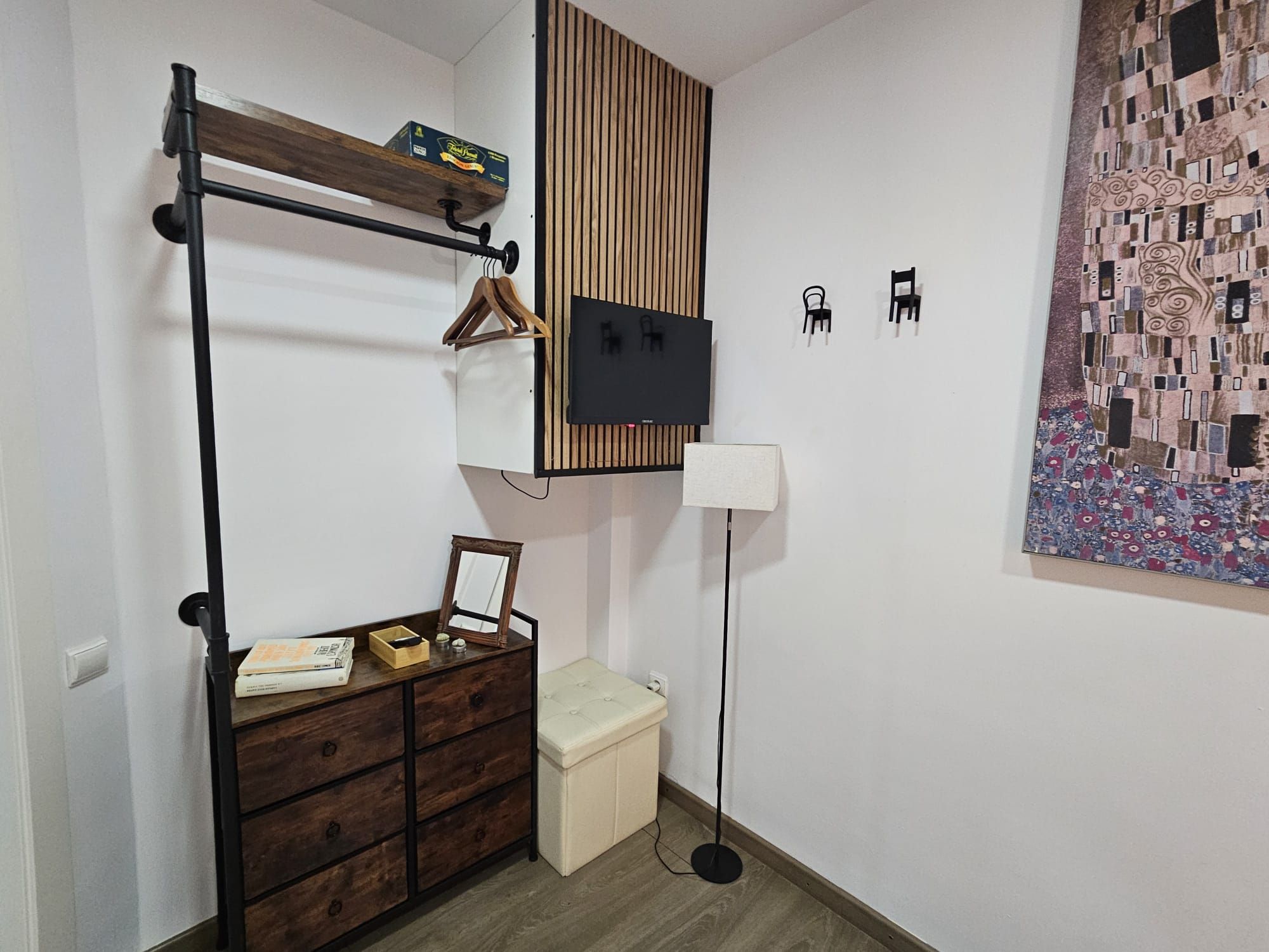 A compact room with a built-in wardrobe, a small bedside table, a floor lamp, and a decorative wall piece.