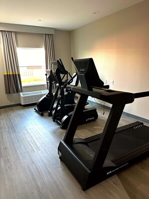 Ein Fitnessraum mit Laufbändern und Crosstrainern, ein großes Fenster mit Vorhängen und ein Holzboden.