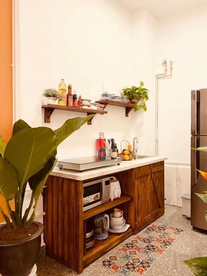 A compact kitchen with wooden cabinets, a microwave, and a countertop with various items. There are two wooden shelves above the counter, and a potted plant is visible. - Villa Premier Village Da Nang Abogo (Ha Noi)