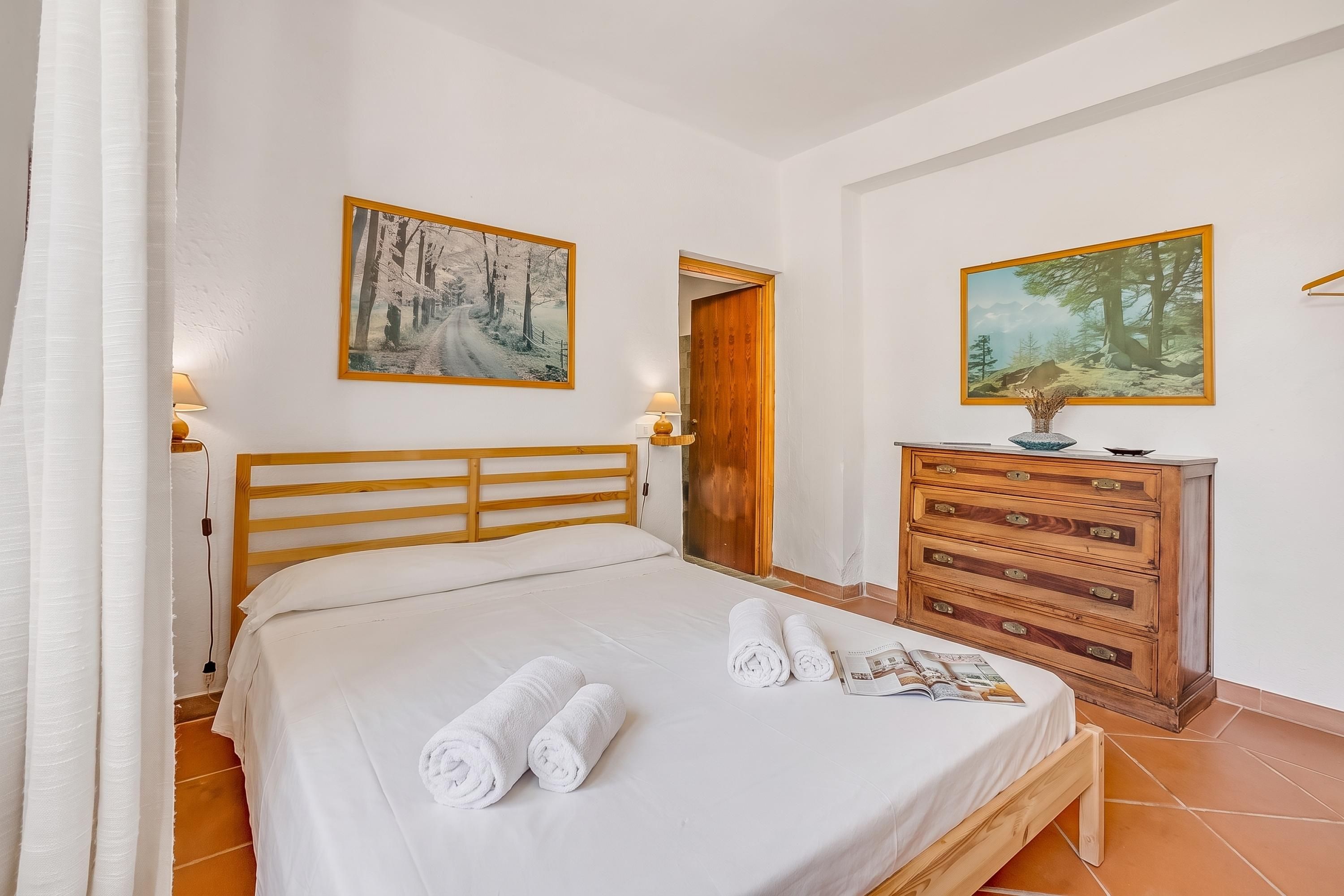 A bedroom with a wooden bed, two paintings on the wall, a wooden dresser, and a door leading to another room.