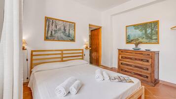 A bedroom with a wooden bed, two paintings on the wall, a wooden dresser, and a door leading to another room.