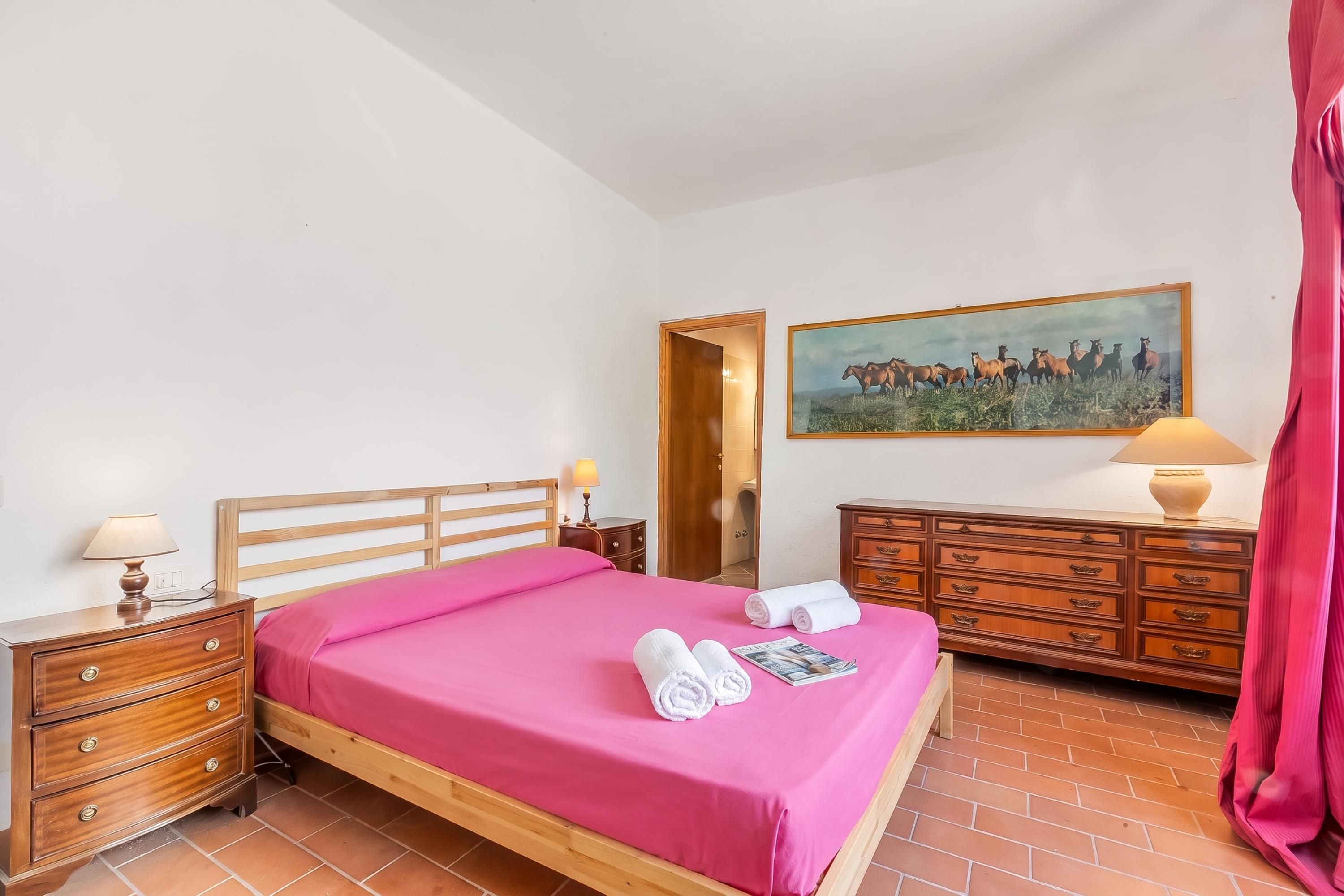 A bedroom with a wooden bed, a nightstand with a lamp, a dresser, a painting on the wall, and a red curtain.