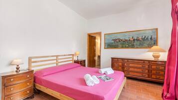 A bedroom with a wooden bed, a nightstand with a lamp, a dresser, a painting on the wall, and a red curtain.