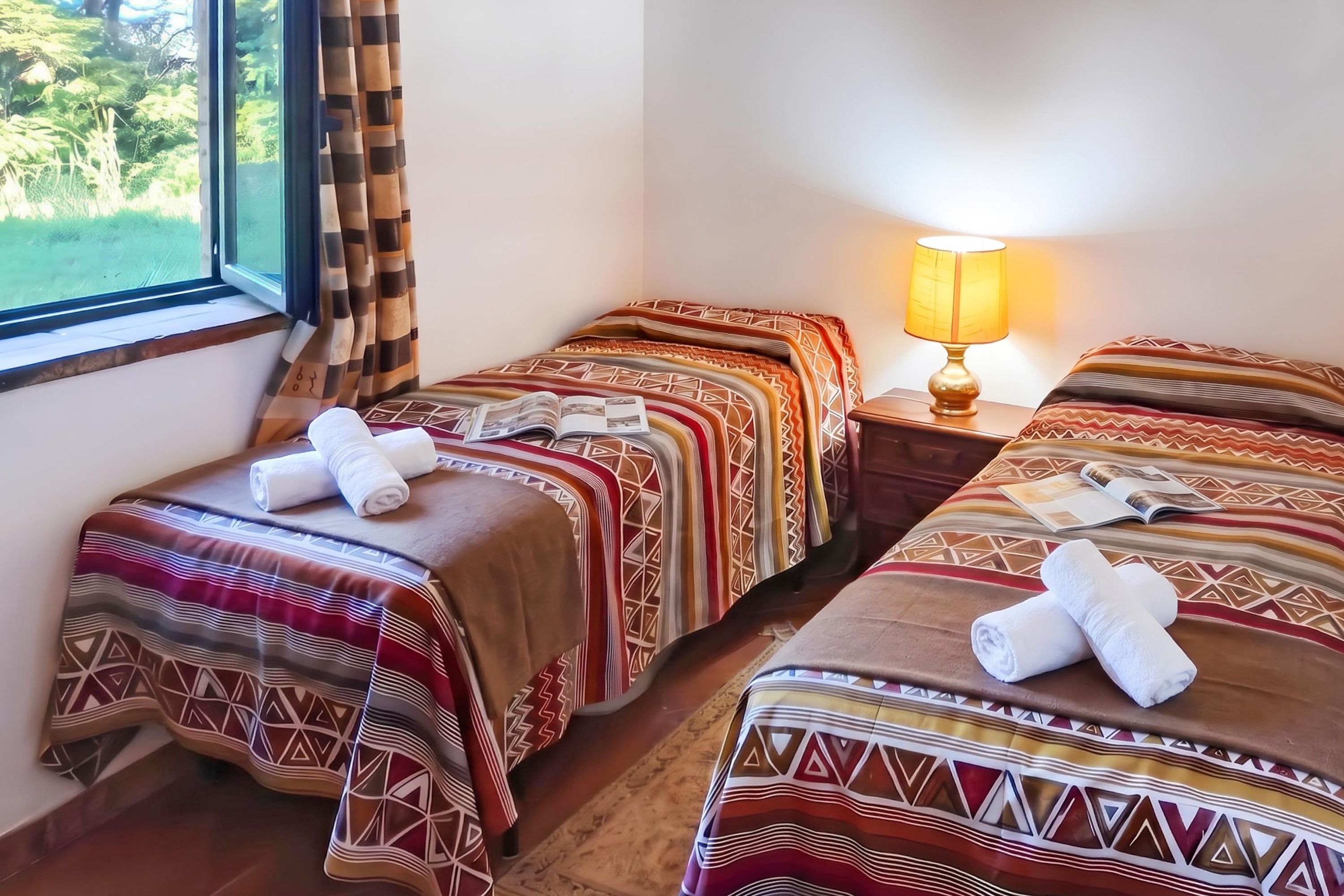 Ein Hotelzimmer mit zwei Betten, jeweils mit gemusterten Bettdecken und Handtüchern, einer Nachttischlampe und einem fensterbepflanzten Fenster mit Vorhängen.