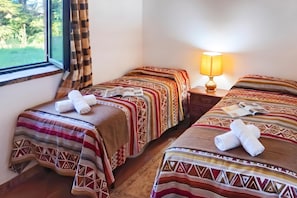 Quarto de hotel com duas camas, cada uma com roupa de cama e toalhas estampadas, uma luminária de cabeceira e uma janela com cortinas.