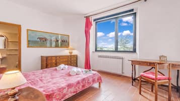 Un dormitorio con cama, escritorio, silla, cómoda, espejo, ventana con vistas y un cuadro en la pared.