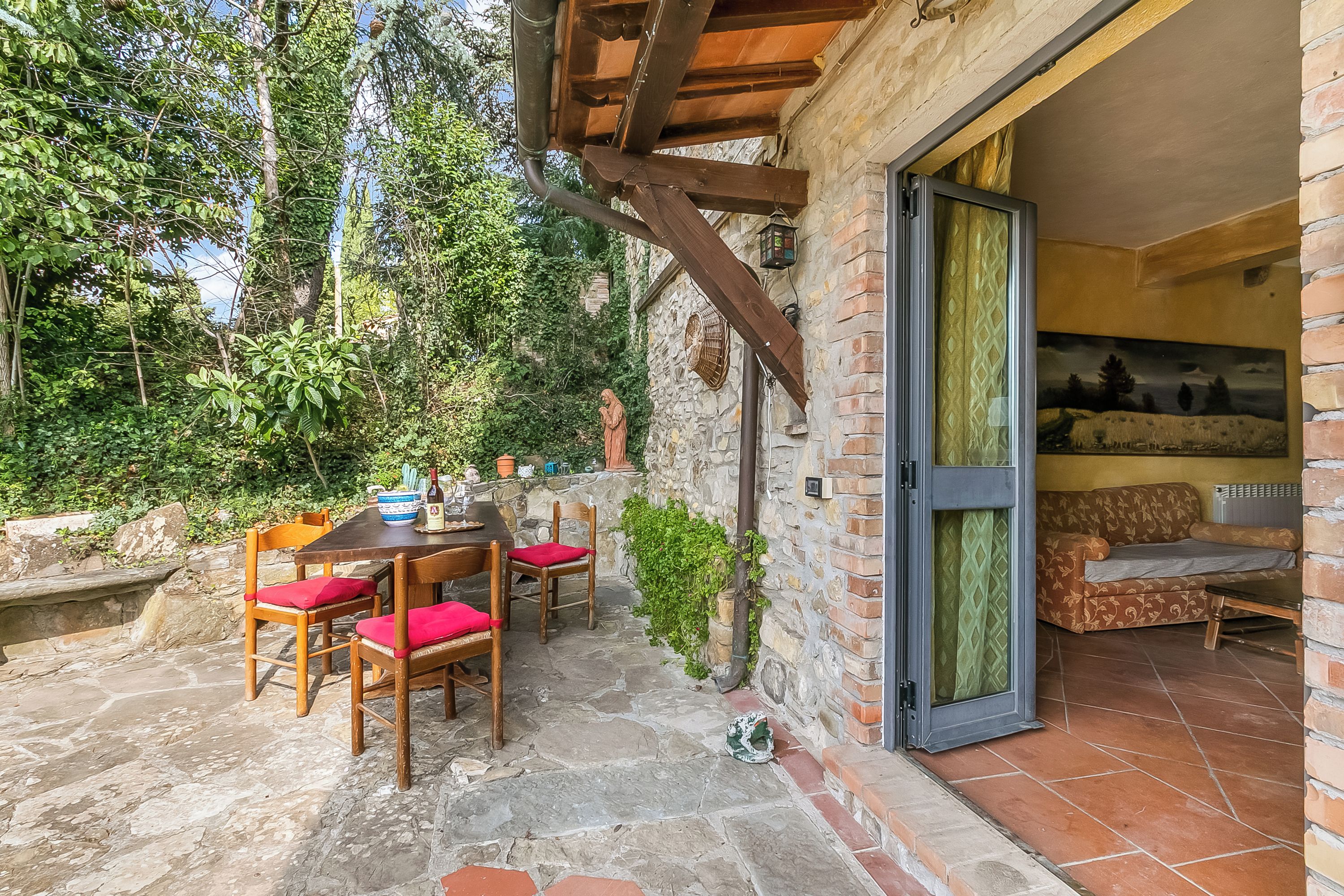 An outdoor patio with a stone table and wooden chairs, a statue, and a view of the garden.