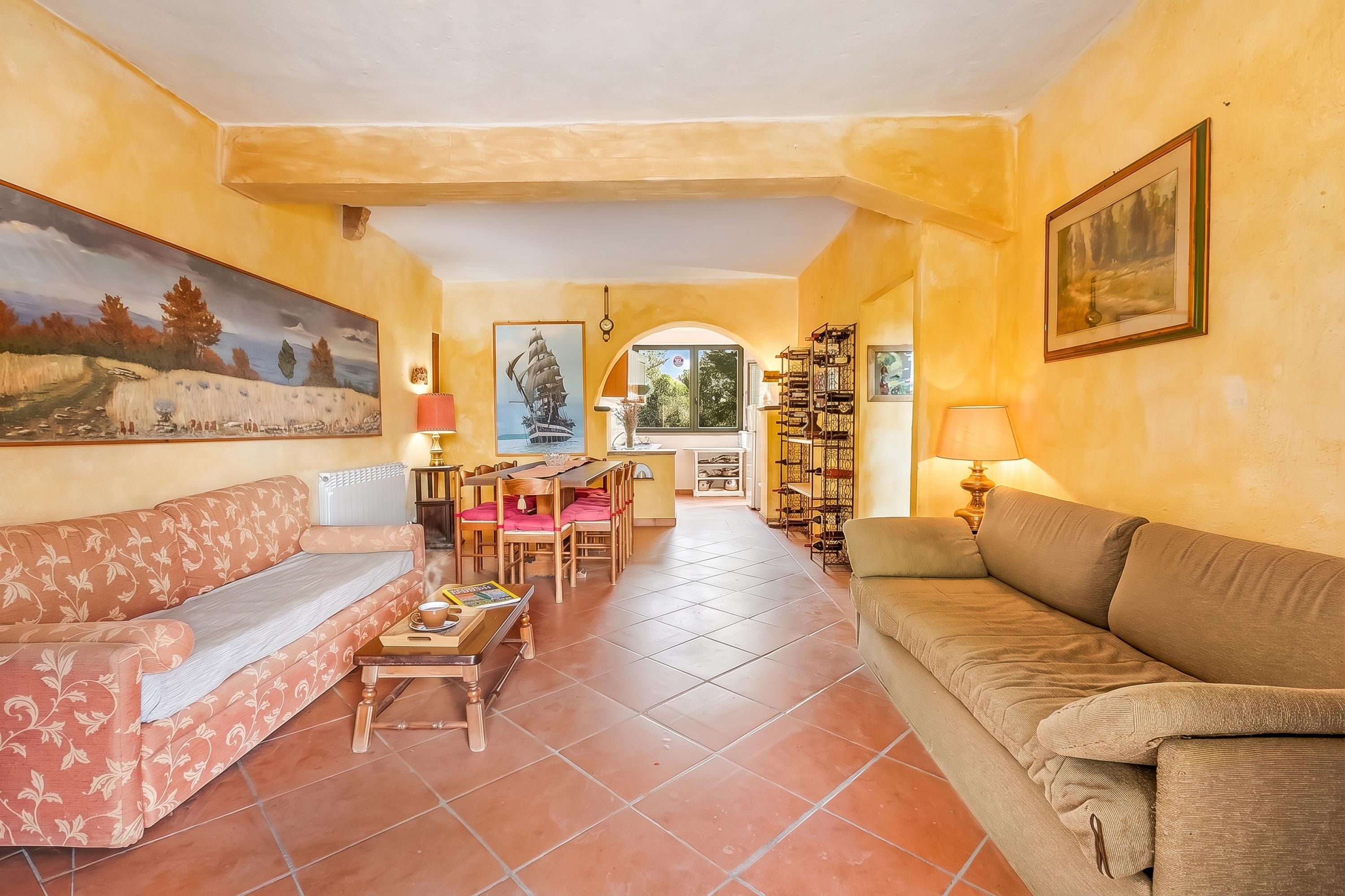 A living room with a sofa, a coffee table, and a dining table with chairs. There are paintings on the walls and a window with a view.